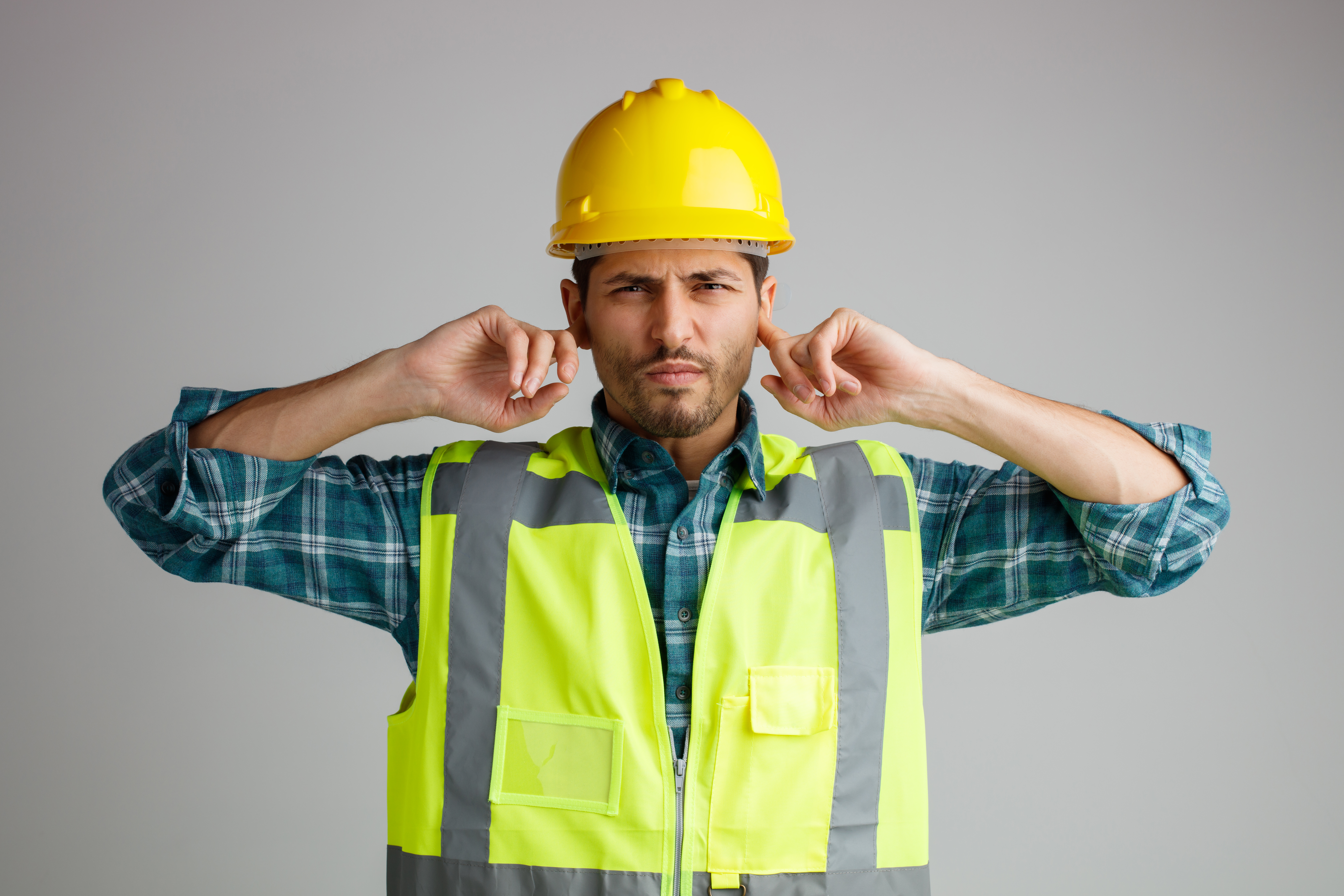 Home unpleased young male engineer wearing safety helmet and uniform looking at camera while keeping fingers in ears isolated on white background